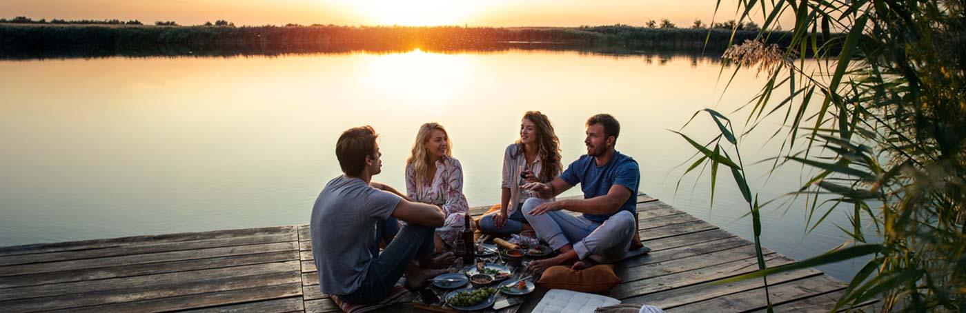 A group of friends sit on the dock of a lake, enjoying a picnic at sunset